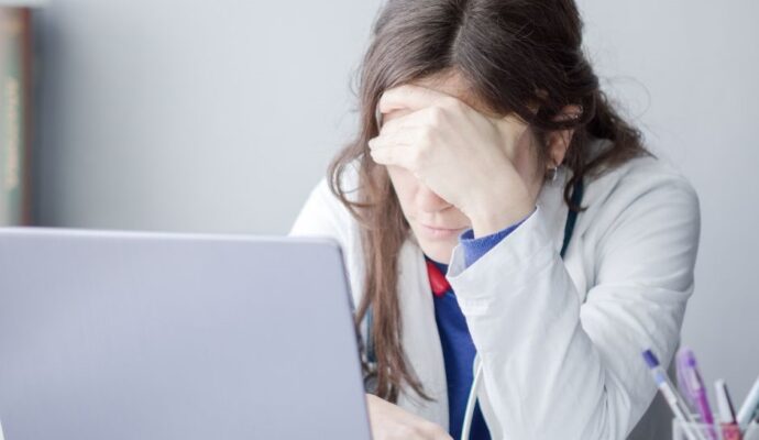 woman doctor with hands on head over a laptop looking tired © Mr KornFlakes via canva.com and deposit photos